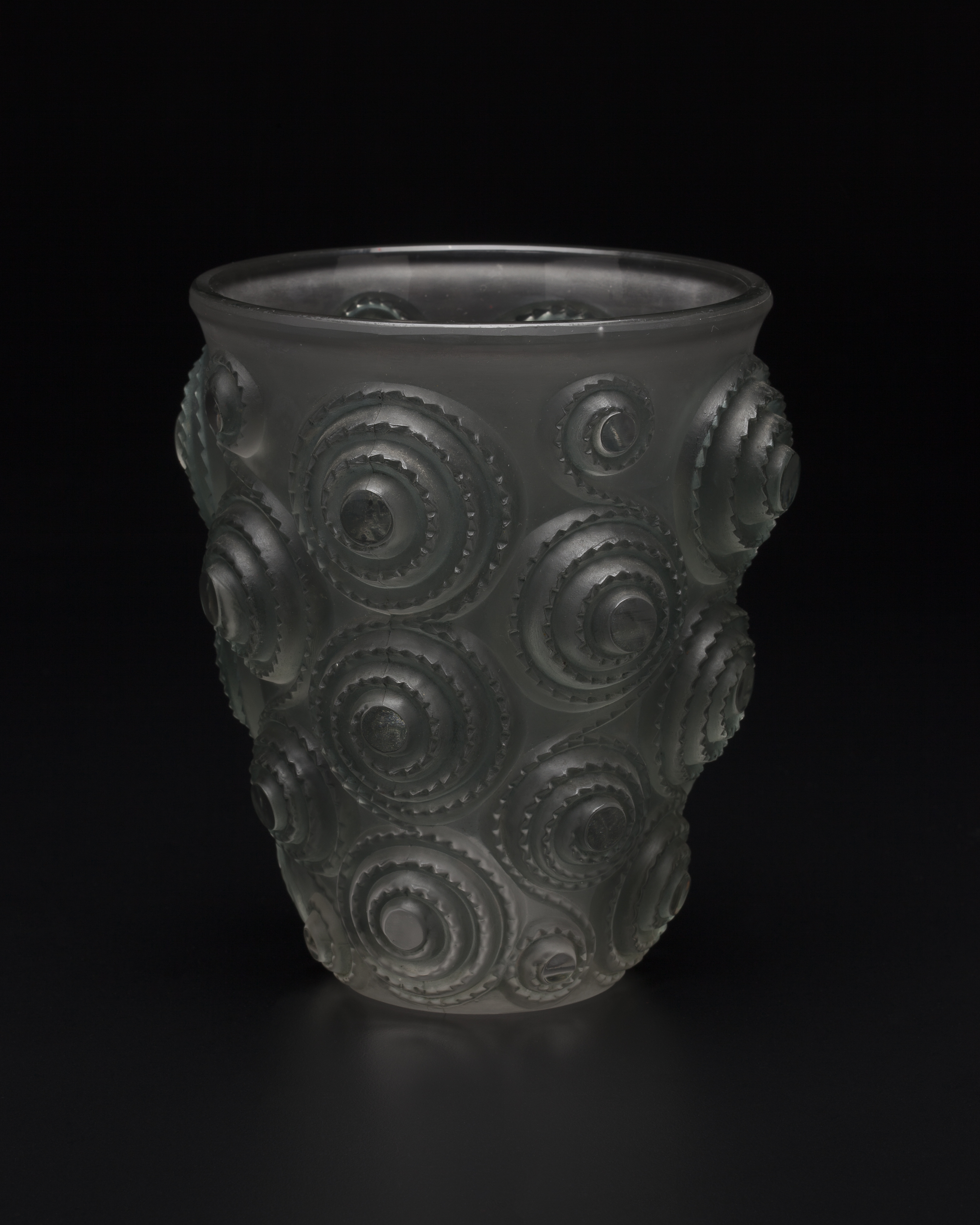 A heavy, cylindrical vase in grey glass with frosted finish, tapering towards the base from a wider rim. The bold, high-relief surface has alternating vertical rows of interlinked notched spiral-shaped forms. 
